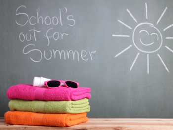 School's out for Summer is written in chalk on a blackboard. In the foreground a pile of beach towels and a pair of sunglasses are piled on a desk.