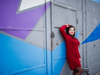a woman in a red woollen dress stands against a geometrically patterned wall.