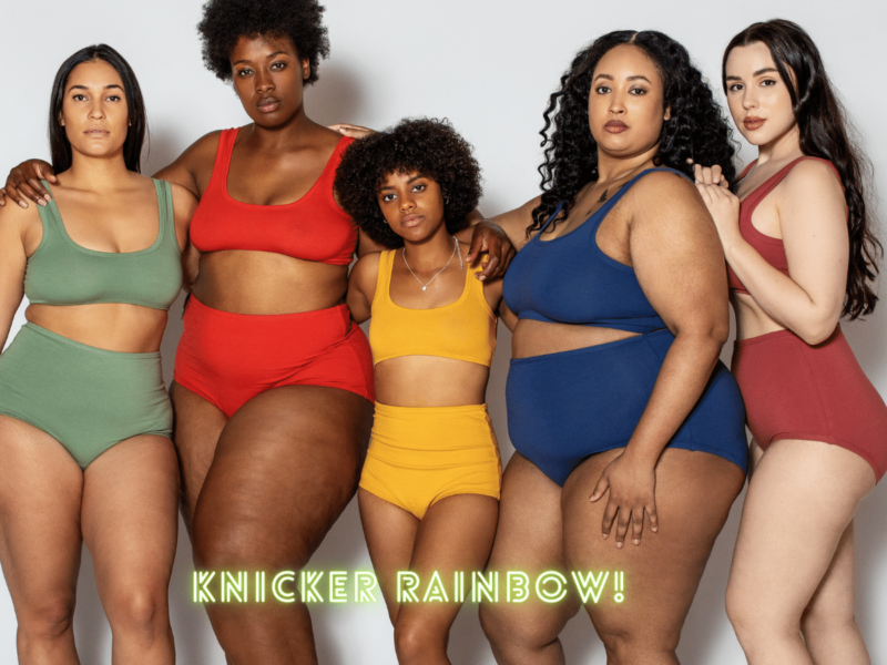 five women stand facing the camera with their arms around each other. They wear bras and knickers in different colours: green, red, yellow, blue and maroon.
