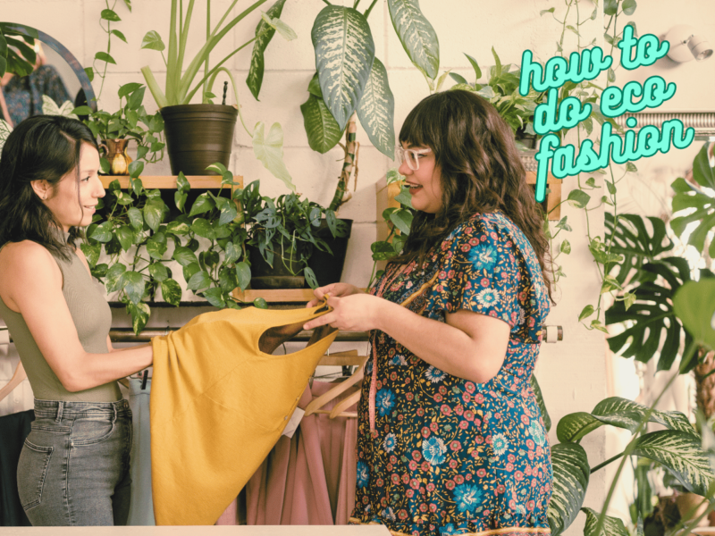 A female clothes maker holds out a mustard coloured top to another woman who is feeling the fabric. There is a rail of clothes in the background and several indoor plants on shelving. Text reads "how to do eco fashion".