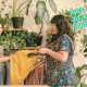 A female clothes maker holds out a mustard coloured top to another woman who is feeling the fabric. There is a rail of clothes in the background and several indoor plants on shelving. Text reads "how to do eco fashion".
